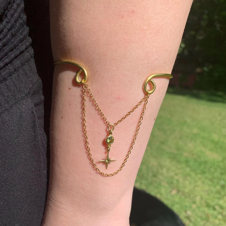 May include: A gold arm cuff with a dangling chain and a star charm with a green gemstone. The cuff is worn on a person's arm.