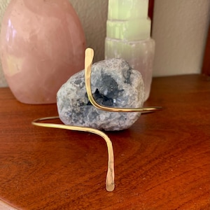 May include: A gold-toned metal headband with a simple, curved design. The headband is shown on a wooden surface with a rock in the background.