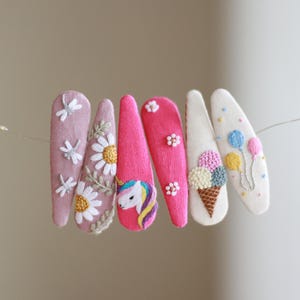 May include: A collection of six embroidered hair clips in various designs. The clips feature a dragonfly, daisies, a unicorn, and ice cream cone, and balloons. The clips are in shades of pink and white.