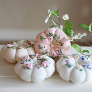 May include: A collection of handmade pincushions in various colors and floral embroidery designs. The pincushions are shaped like pumpkins and feature detailed stitching with a pearl accent. The color palette includes soft pinks, whites, and a variety of floral hues.