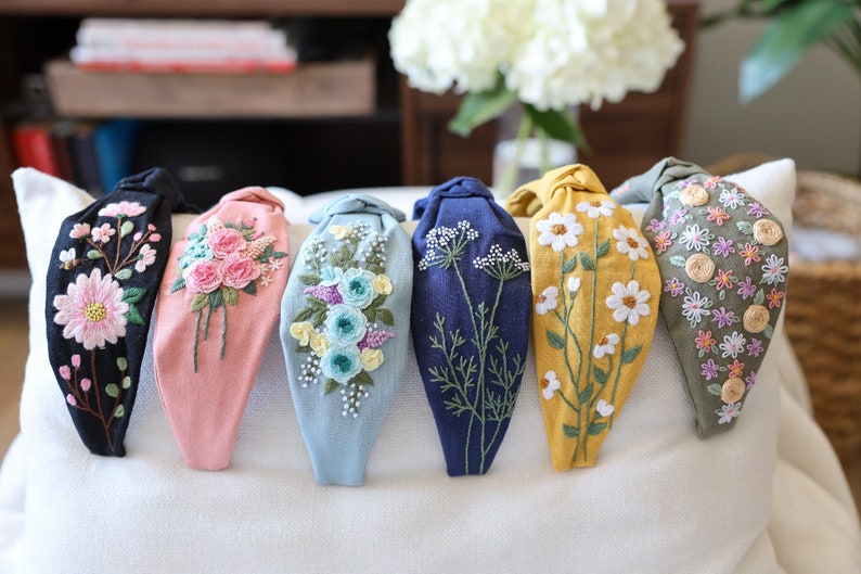 Pode incluir: Uma cole&ccedil;&atilde;o de seis tiaras de tecido, cada uma adornada com bordados florais intrincados. As tiaras apresentam uma variedade de cores, incluindo preto, rosa, azul, azul marinho, amarelo e verde oliva. As flores bordadas incluem margaridas, rosas e outras flores delicadas.