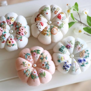 May include: Four floral-shaped pin cushions in white and pink. Each cushion features detailed embroidery of flowers and foliage in various colors. A pearl bead is centered on each cushion.