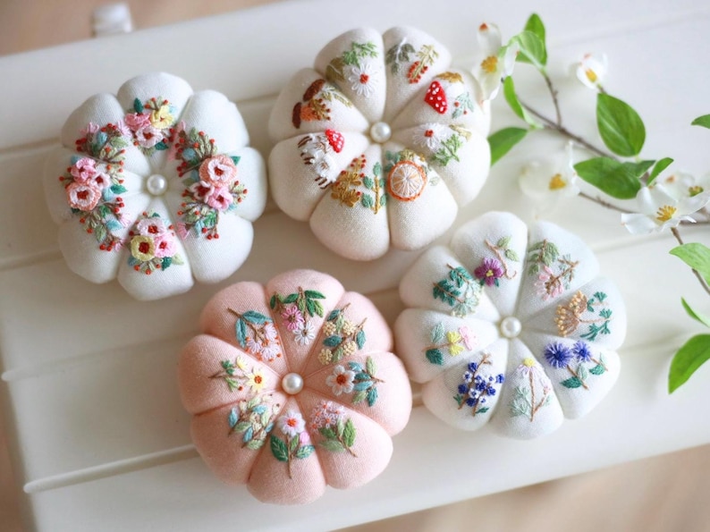 May include: Four floral-shaped pin cushions in white and pink. Each cushion features detailed embroidery of flowers and foliage in various colors. A pearl bead is centered on each cushion.