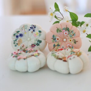 May include: Four handmade pincushions in a flower shape, each with embroidered floral designs. Two are white, one is peach, and all have a pearl center. The pincushions are arranged on a white surface, with a blurred floral background.