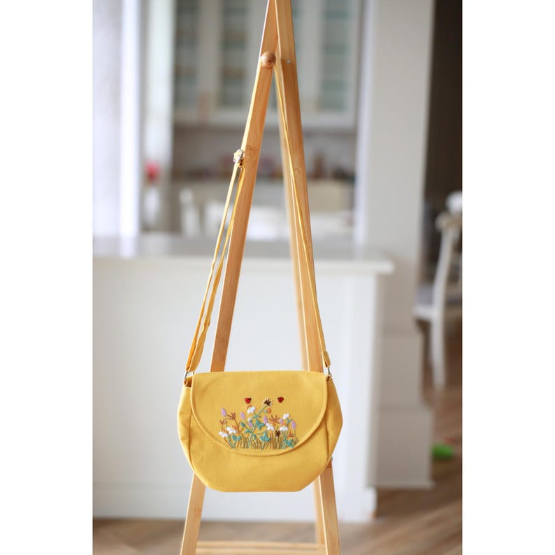 May include: Mustard yellow crossbody bag with a flap closure. The bag features an embroidered floral design in white, red, and green. The bag is hanging on a wooden A-frame stand.