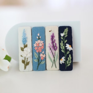 May include: Four embroidered hair clips with floral designs. Each clip features a different flower: a blue flower, pink flowers, purple lavender, and white daisies. The clips are on a white background.