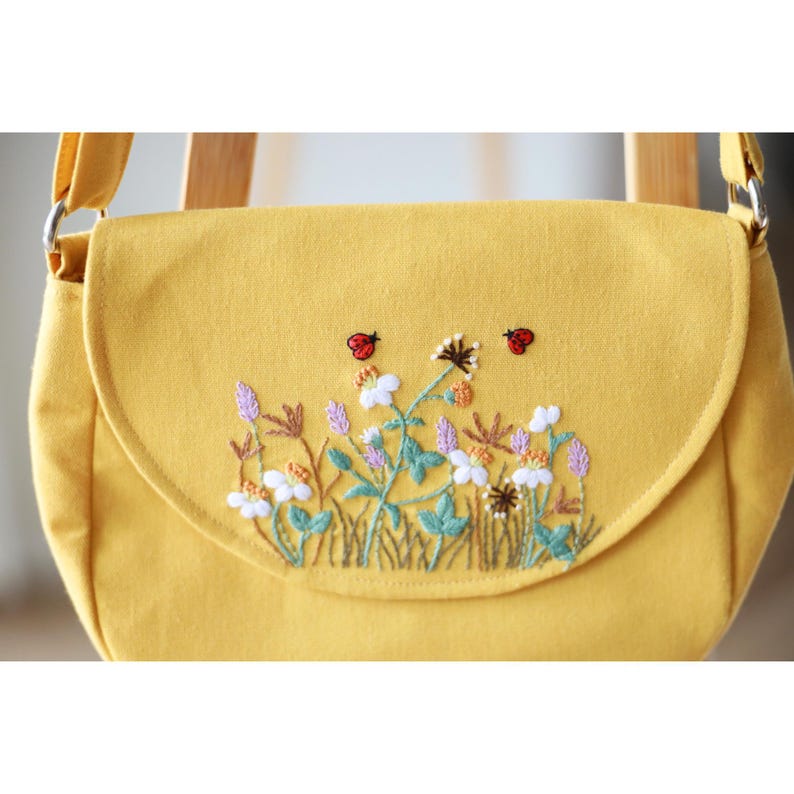 May include: A yellow crossbody bag with a flap closure. The bag features an embroidered design of wildflowers, green grass, and two red ladybugs. The strap and the bag are made of the same yellow fabric.