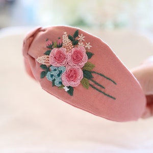 Pode incluir: Uma tiara de tecido rosa com um design floral bordado. O bordado apresenta rosas cor-de-rosa, flores azuis e folhas verdes. A tiara &eacute; um acess&oacute;rio elegante.