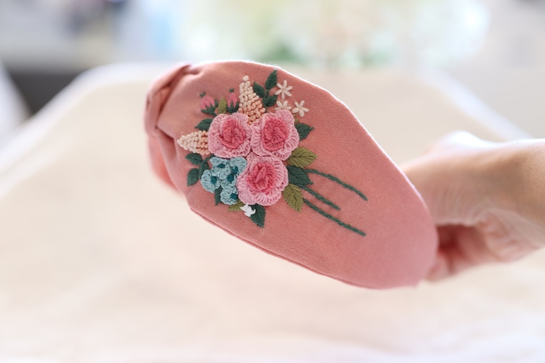 Pode incluir: Uma tiara de tecido rosa com um design floral bordado. O bordado apresenta rosas cor-de-rosa, flores azuis e folhas verdes. A tiara &eacute; um acess&oacute;rio elegante.