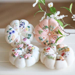 May include: Four handmade pincushions in a variety of colors. Two are white, one is pink. Each pincushion is round with a petal-like design and embroidered floral patterns. A pearl is in the center of each pincushion.