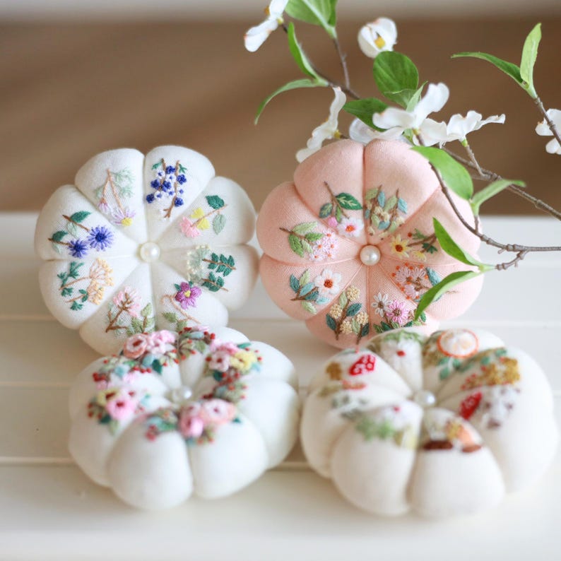 May include: Four handmade pincushions in a variety of colors. Two are white, one is pink. Each pincushion is round with a petal-like design and embroidered floral patterns. A pearl is in the center of each pincushion.