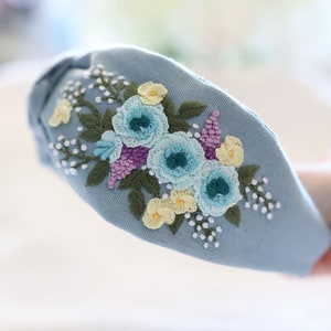 Pode incluir: Uma tiara azul clara com flores bordadas em tons de azul, amarelo e roxo. O design floral &eacute; detalhado com folhas verdes e detalhes brancos, criando um acess&oacute;rio delicado e feminino.