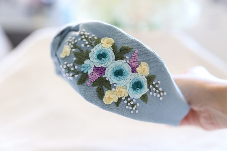 Pode incluir: Uma tiara azul clara com flores bordadas em tons de azul, amarelo e roxo. O design floral &eacute; detalhado com folhas verdes e detalhes brancos, criando um acess&oacute;rio delicado e feminino.