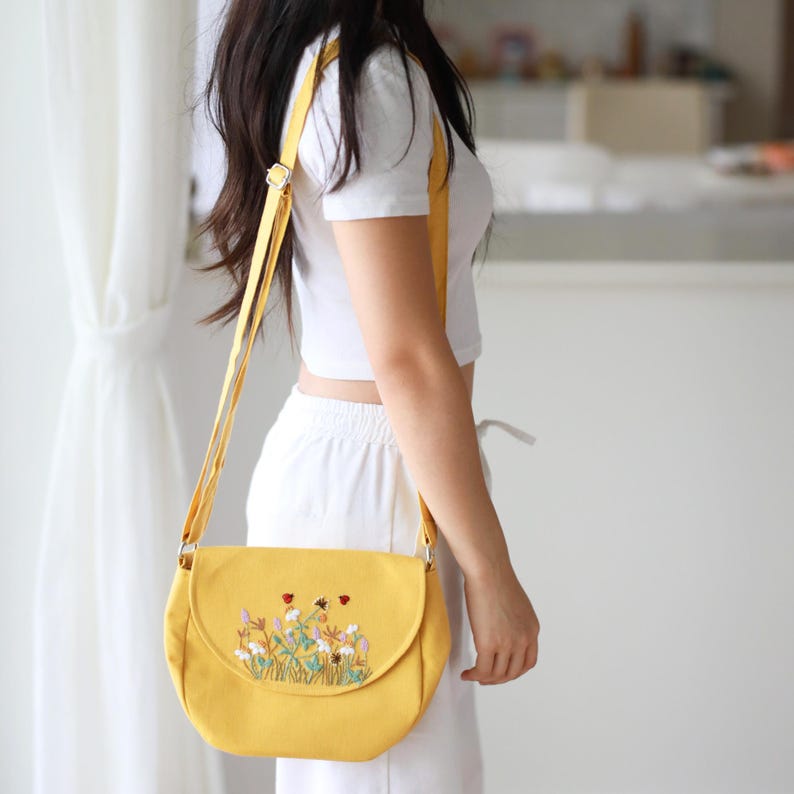 May include: A yellow crossbody bag with a flap closure and adjustable strap. The bag features an embroidered floral design with red ladybugs and various flowers. The bag is made of a soft material.