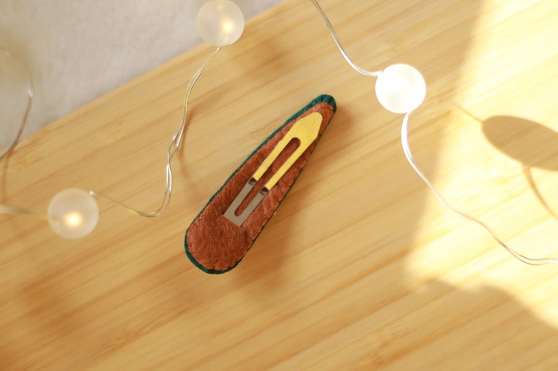 K&ouml;nnte beinhalten: Eine handgefertigte Haarspange mit einem goldfarbenen Metallverschluss. Die Spange ist mit braunem Filz mit einem gr&uuml;nen Rand bedeckt. Die Spange liegt auf einer Holzoberfl&auml;che, mit Lichterketten im Hintergrund.