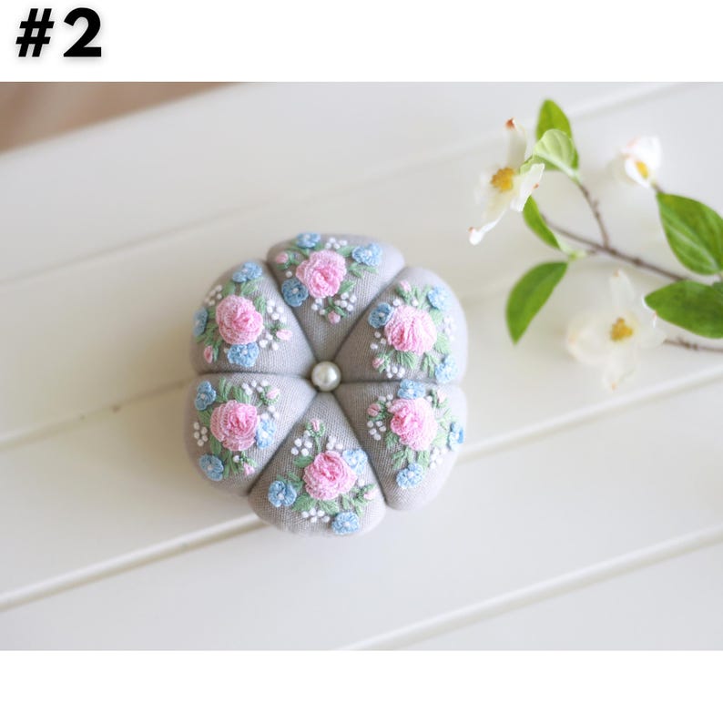 Pu&ograve; includere: Un cuscino per spilli in tessuto grigio con un bottone di perla al centro. Il cuscino &egrave; ricamato con fiori rosa e blu.