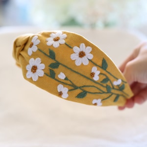 Pode incluir: Tiara amarelo mostarda com bordado de margaridas brancas e caules verdes. O desenho floral &eacute; costurado &agrave; m&atilde;o na parte superior da tiara. Um acess&oacute;rio de cabelo elegante para uso di&aacute;rio.