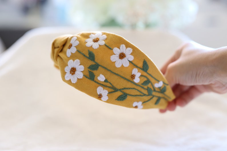 Pode incluir: Tiara amarelo mostarda com bordado de margaridas brancas e caules verdes. O desenho floral &eacute; costurado &agrave; m&atilde;o na parte superior da tiara. Um acess&oacute;rio de cabelo elegante para uso di&aacute;rio.