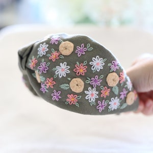 Pode incluir: Tiara verde-oliva com design floral bordado. A tiara apresenta um top amarrado e &eacute; adornada com flores brancas, rosas e roxas, juntamente com pequenos detalhes circulares bege e folhas verdes.