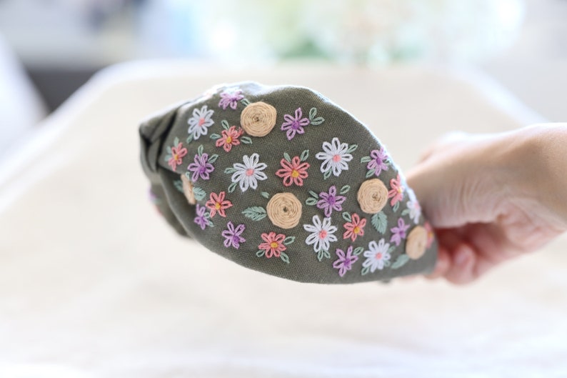 Pode incluir: Tiara verde-oliva com design floral bordado. A tiara apresenta um top amarrado e &eacute; adornada com flores brancas, rosas e roxas, juntamente com pequenos detalhes circulares bege e folhas verdes.