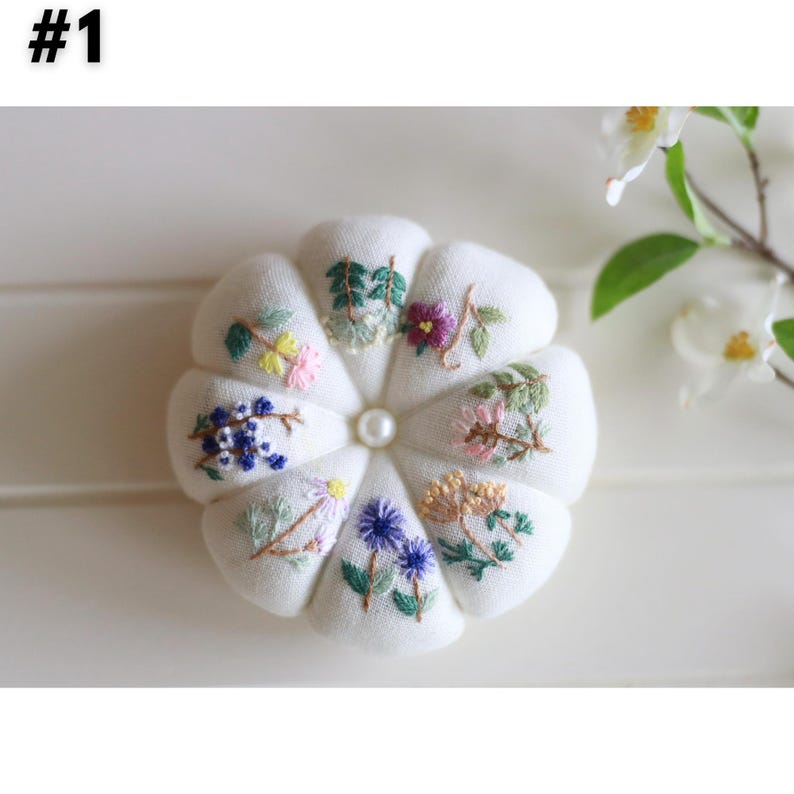 May include: A white, flower-shaped pin cushion with embroidered floral designs in various colors. The cushion has a pearl-like center and is surrounded by detailed, colorful floral embroidery. The image is well-lit, highlighting the texture and detail of the embroidery.