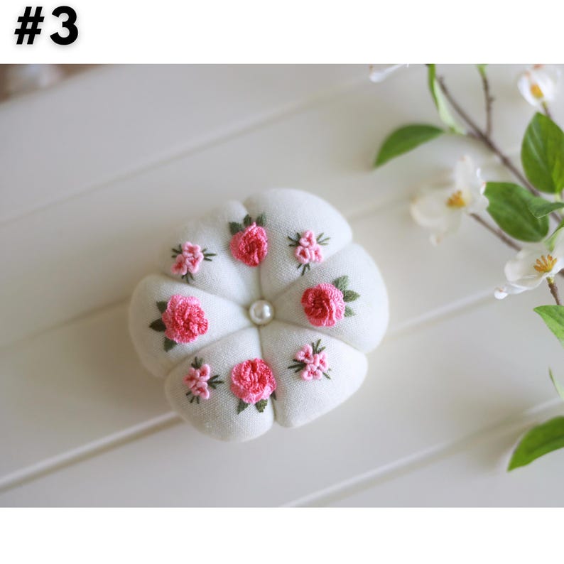 Pu&ograve; includere: Un cuscino per spilli in tessuto bianco con un bottone di perla al centro e rose rosa e foglie verdi ricamate.