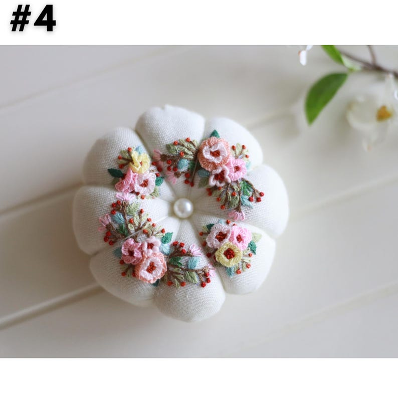 May include: A white, flower-shaped pin cushion with embroidered floral accents in pink, yellow, and green. A pearl bead is centered in the design. The number "#4" is in the upper left corner.