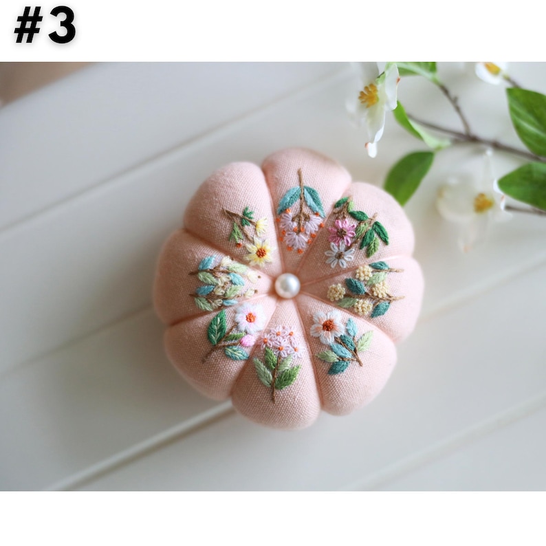 May include: A round, light pink pincushion with embroidered floral designs in various colors. The pincushion has a central pearl bead and is shaped like a pumpkin. The number "#3" is in the top left corner.