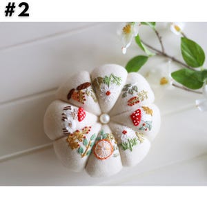 May include: Cream-colored, round pin cushion with embroidered floral and mushroom designs. The cushion has a central pearl bead and is surrounded by white flowers and green leaves. The image has the number "#2" in the top left corner.