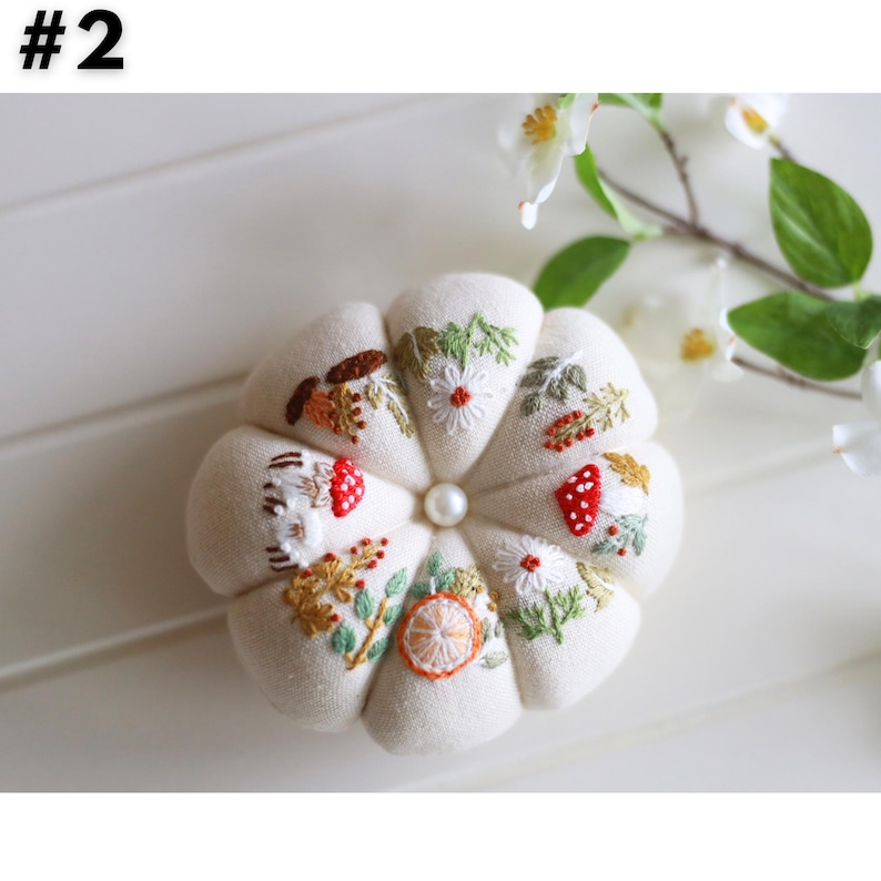 May include: Cream-colored, round pin cushion with embroidered floral and mushroom designs. The cushion has a central pearl bead and is surrounded by white flowers and green leaves. The image has the number "#2" in the top left corner.