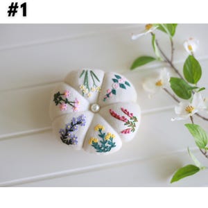 Pu&ograve; includere: Un cuscino per spilli bianco con un bottone di perla al centro. Il cuscino ha la forma di un fiore e ha sei petali, ognuno ricamato con un diverso tipo di fiore in vari colori.