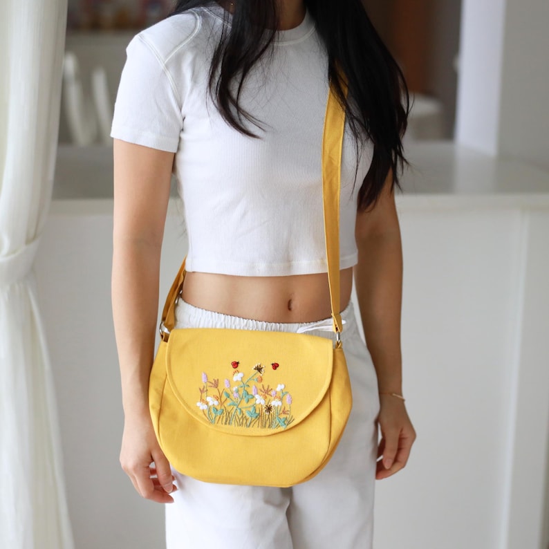 May include: A yellow crossbody bag with a flap closure and a floral embroidery design. The bag has a matching yellow strap and is made of a sturdy fabric. The embroidery features colorful flowers and insects.