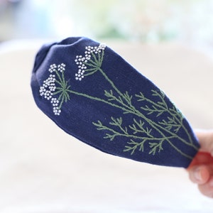 Pode incluir: Uma faixa de cabelo azul marinho com bordados de cachos de flores brancas e caules verdes. O desenho floral est&aacute; no lado esquerdo da faixa de cabelo. A faixa de cabelo &eacute; segurada por uma m&atilde;o.