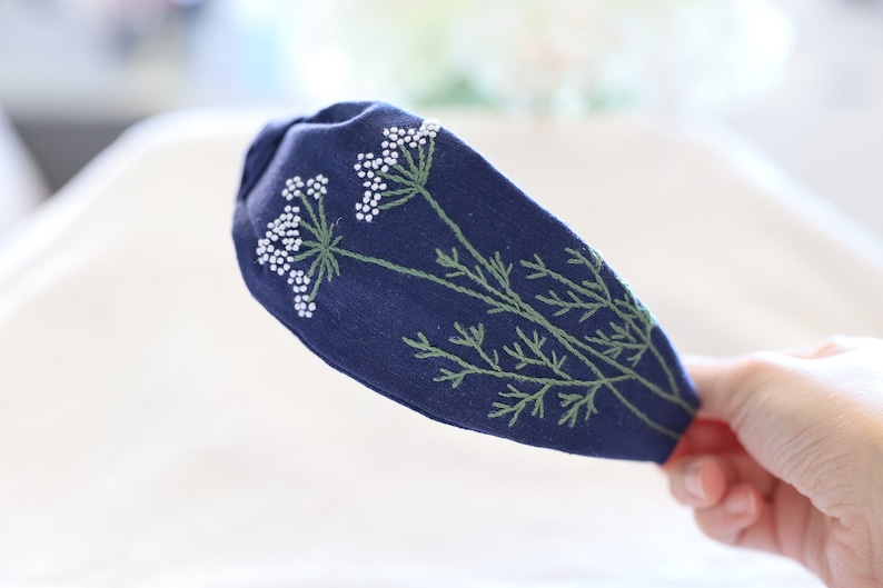 Pode incluir: Uma faixa de cabelo azul marinho com bordados de cachos de flores brancas e caules verdes. O desenho floral est&aacute; no lado esquerdo da faixa de cabelo. A faixa de cabelo &eacute; segurada por uma m&atilde;o.