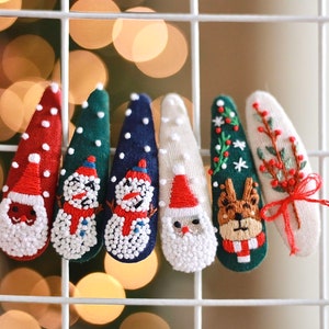 K&ouml;nnte beinhalten: Eine Sammlung von Haarspangen mit Weihnachtsmotiven. Jede Spange zeigt gestickte Designs, darunter der Weihnachtsmann, Schneem&auml;nner, ein Rentier und festliches Gr&uuml;n. Die Spangen sind in verschiedenen Farben gehalten, darunter Rot, Gr&uuml;n und Blau, mit wei&szlig;en Akzenten.
