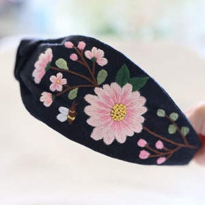 Pode incluir: Uma faixa de cabelo azul marinho com flores rosa bordadas, folhas verdes e uma abelha. O design floral &eacute; centrado na faixa, criando um acess&oacute;rio decorativo.