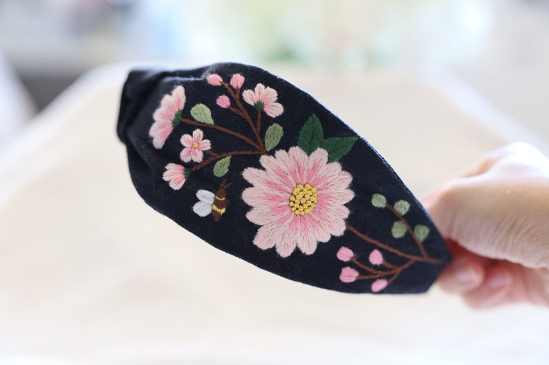 Pode incluir: Uma faixa de cabelo azul marinho com flores rosa bordadas, folhas verdes e uma abelha. O design floral &eacute; centrado na faixa, criando um acess&oacute;rio decorativo.