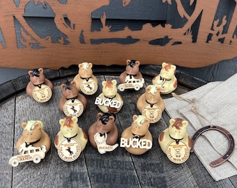 May include: A collection of 10 brown and tan rubber duckies with horse heads and wooden tags that say "You've Been Bucked".  Some of the ducks have a wooden tag that says "Bucked" and one duck has a wooden tag that says "You've Been Bucked" and a small wooden toy truck.  The ducks are sitting on a wooden barrel and a brown burlap bag with a horseshoe.