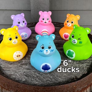 May include: Six colorful rubber duck toys shaped like bears, each with unique facial expressions and designs. The ducks are approximately 6 inches in size and are arranged on a wooden barrel.