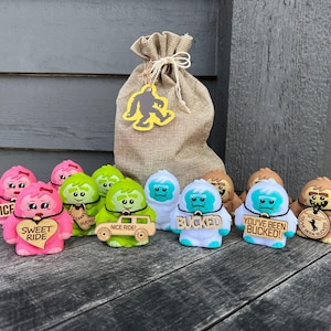 May include: A set of nine plush toys shaped like snowmen with different colored faces and accessories. The toys are arranged in a row on a wooden surface. The toys have different sayings on them, including "Nice Ride", "Sweet Ride", "You've Been Bucked!" and "Bucked".