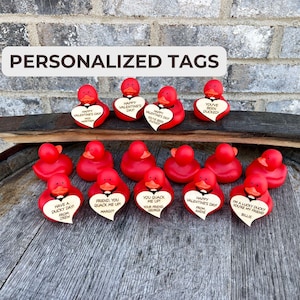 May include: A collection of red rubber duckies with personalized Valentine's Day messages on heart-shaped tags. The ducks are arranged on a wooden barrel.