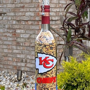 May include: A glass wine bottle bird feeder with a red top and a white sticker featuring the Kansas City Chiefs logo. The bottle is filled with birdseed and is suspended from a chain.