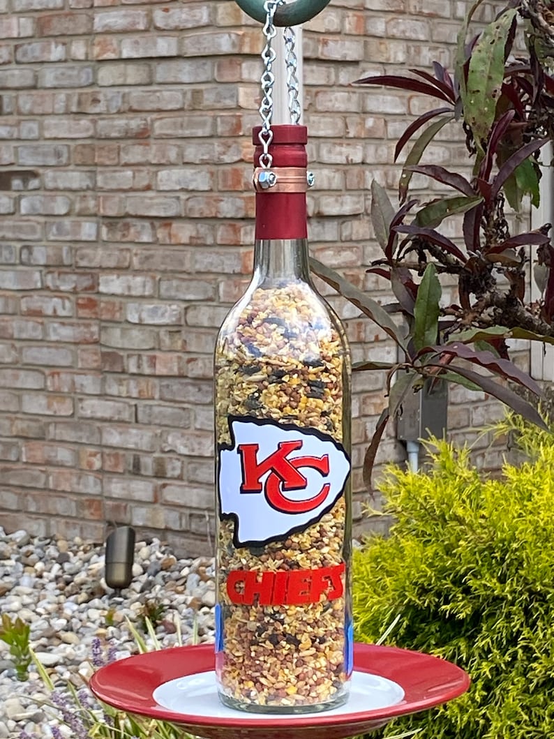 May include: A glass wine bottle bird feeder with a red top and a white sticker featuring the Kansas City Chiefs logo. The bottle is filled with birdseed and is suspended from a chain.