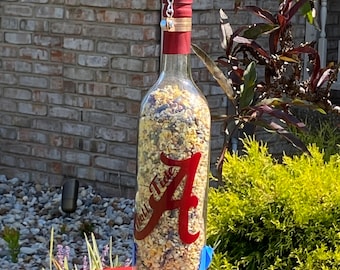 Könnte beinhalten: Ein Vogelfutterspender aus einer klaren Glasflasche gefüllt mit Vogelfutter, mit einem roten Schriftzug Roll Tide und einem großen roten Buchstaben A. Die Flasche hängt an einer silbernen Kette und ruht auf einem runden, rot-weißen Keramikt-weißen Teller als Basis.