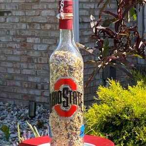 May include: A glass bottle bird feeder with a red top and a chain hanging from it. The bottle is filled with birdseed and has a black and red Ohio State logo on it. The feeder is hanging over a red saucer.