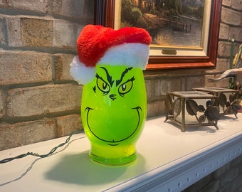 May include: A green glass Christmas decoration with the Grinch's face, wearing a red and white Santa hat. The decoration is lit, casting a shadow on the white mantelpiece. A painting hangs in the background.