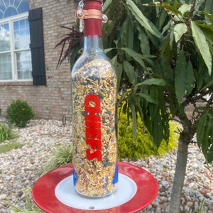 May include: A red and white bird feeder made from a glass wine bottle with a red label that says "Cheers". The feeder is hanging from a chain and has a red saucer underneath.