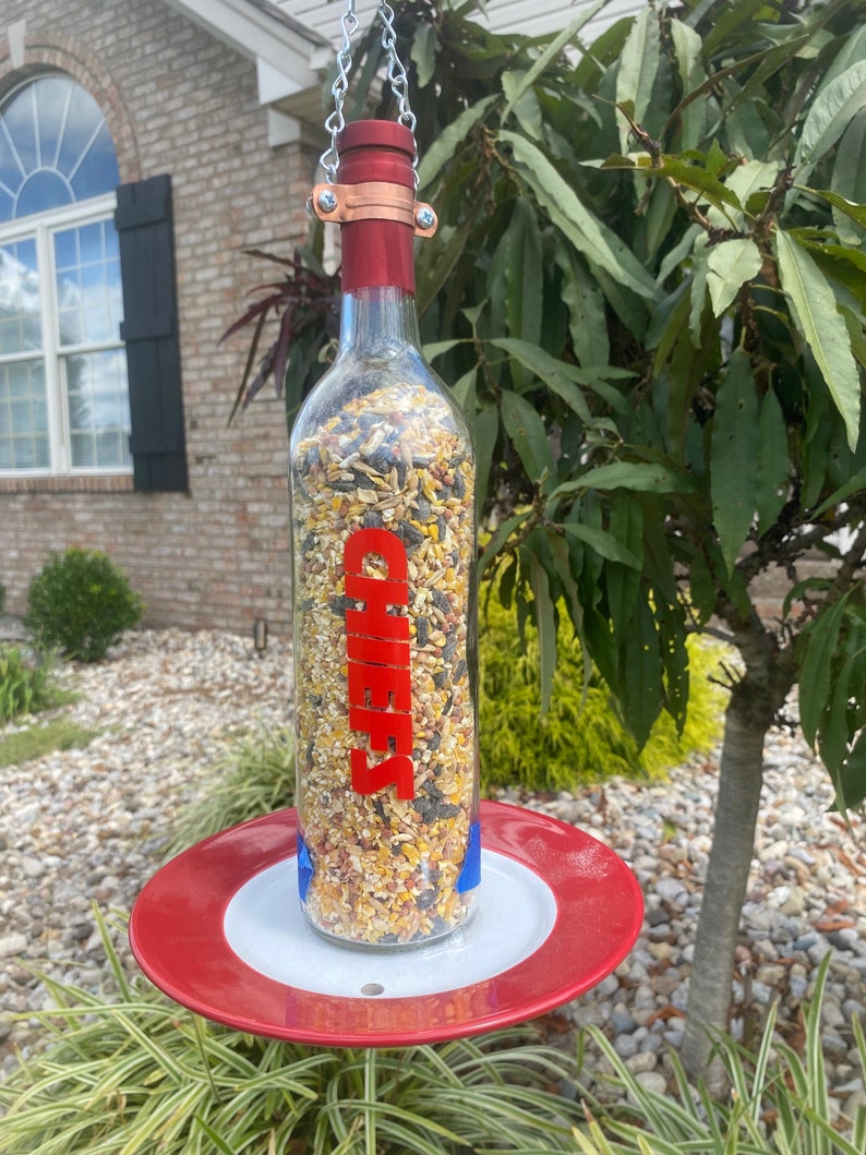 May include: A red and white bird feeder made from a glass wine bottle with a red label that says "Cheers". The feeder is hanging from a chain and has a red saucer underneath.