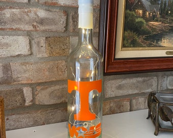 Könnte beinhalten: Eine klare Glasflasche mit weißem Verschluss, beleuchtet von Lichterketten. Die Flasche hat ein orangefarbenes "T" und das "Vol"-Logo. Die Flasche ist ein dekorativer Gegenstand, möglicherweise eine Lampe oder ein Mittelstück.