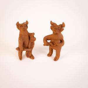 May include: Two terracotta figurines of bull-like creatures. One is holding a long, thin object, while the other is holding a round object.